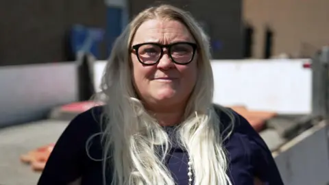 Katy Bald is smiling at the camera. She has long, light-coloured hair and is wearing dark-framed glasses, a dark top, and a long beaded necklace.