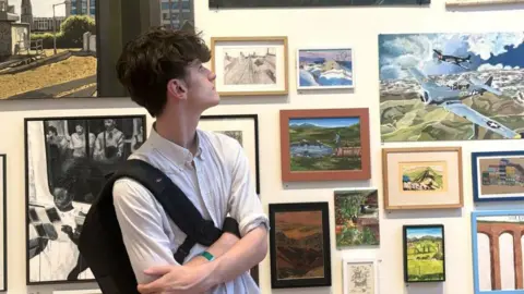 A man with brown hair stands looking up at one particular painting on a wall filled with them. 