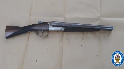 West Midlands Police A brown and silver shotgun which has been laid down on some light brown material.