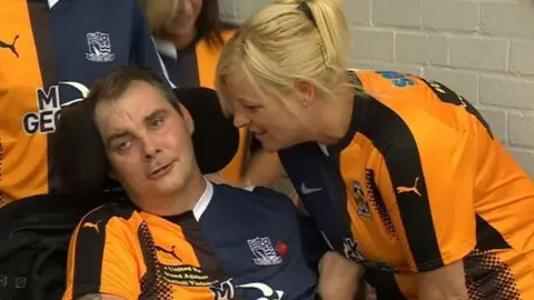 Simon Dobbin in a wheelchair, sitting next to Nicole Dobbin. They are both wearing half-and-half shirts. One half is the orange and black Cambridge United strip, the other is the dark blue Southend kit.