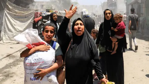 Reuters A woman raises her hand, crying out, as she walks beside a child carrying a sack on a street. a woman behind her carries a small child.