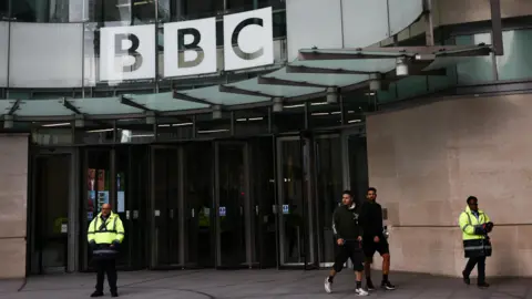 Reuters People walking outside New Broadcasting House - the BBC logo can be seen on a curved glass frontage. Two security guards in high-vis are also standing outside 