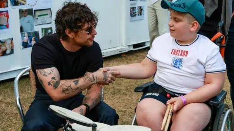 Alex Pope/BBC Boy wearing a green cap and an England football shirt shakes hands with Tom Grennan who is wearing dark glasses. Alfie sits in a wheelchair and holds drum sticks. A drum kit is in front of him. They are backstage and a wall behind has photos on with people milling about. 