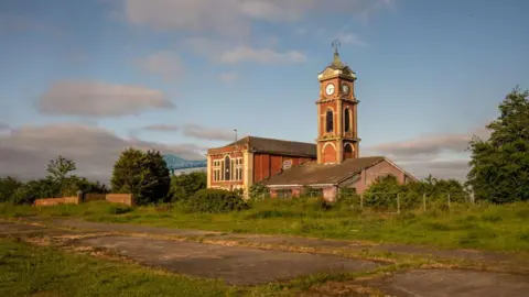 Middlesbrough's derelict Old Town Hall in the Middlehaven area. It is built from red brick and has a tower with a spire. It is surrounded by wasteland. The Tees Transporter Bridge can be seen in the distance.