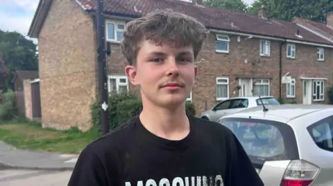 Kate Bradbrook/BBC A young man in a black T-shirt. He is standing in a residential street, in front of a row of terraced houses.