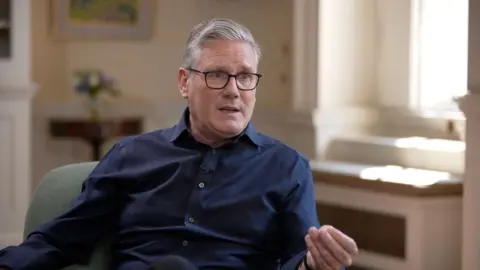 Keir Starmer, with short grey hair and black glasses sits in a chair