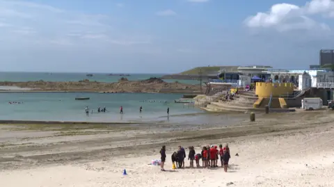 Havre des Pas beach and bathing pool with the sea in the distance. There is a group of people on the beach and some people paddling in the pool. Some people are sitting on the concrete steps leading down from a pavilion in to the pool. 