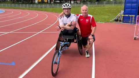 Dr Daniel Gordon Dr Gordon is sitting in a hand bike with his friend. They are posing next to a race track.