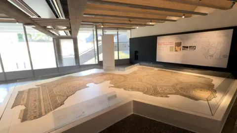 An incomplete Roman mosaic laid out on a white surface with information boards around the side.