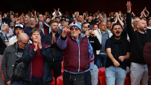 Reuters Aston Villa fans cheering in a stadium. They are wearing claret, blue and sky blue hates, jumpers and scarfs colours of the club