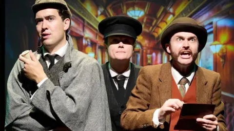 Sheringham Theatre Lewes Roberts is dressed as Sherlock Holmes in a deer-stalker hat, cape and holding a pipe, next to a train conductor wearing a black uniform and peak cap, and a man who is holding a clip board and shouting at the camera. This actor is wearing a brown jacket, waistcoat and tie and brown bowler hat.