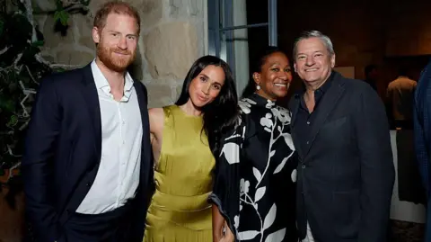 Harry and Meghan smiling next to two others, wearing cocktail attire, at a house
