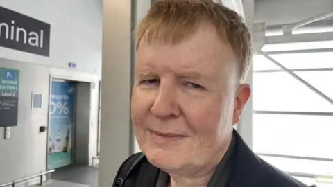 Helen Mulroy/BBC Frank Brown looks at the camera in a gloomy way. He was wearing a shirt, jacket and had a bag strap on his shoulder. It stands in front of some of the terminal sign.