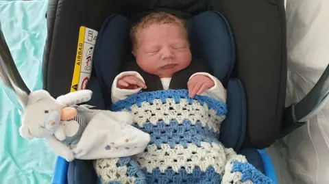Natalie Needham A baby wrapped in a blue and white blanket with a white teddy next to him
