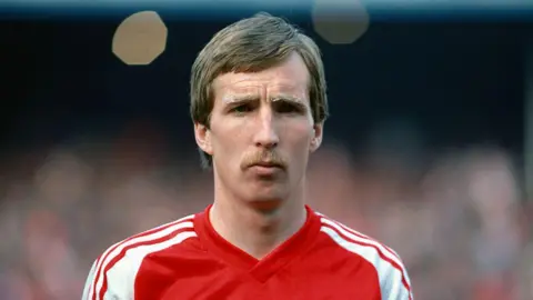 Getty Images A close up of Joey Jones pictured in May 1980 at the British Home Championship - Wales v Northern Ireland. He is staring directly at the camera with a concentrated demeanour and is wearing his red Welsh jersey with white stripes on the shoulders. He has short sandy coloured hair and a moustache. 
