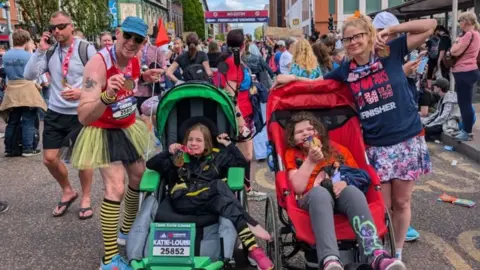 Derek Goodfellow A man in a red vest, yellow and black tutu and striped socks, a hat and sunglasses, posing beside a young girl with brown hair and a black top - in a specialist green run chair. They are pictured beside a woman with long blonde hair and glasses, in a navy top and patterned skirt, who is also beside a young brown haired girl in a specialist red run chair. There are numerous marathon goers in the background.