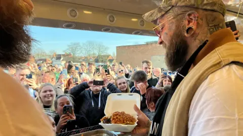 A man with a camouflage cap, face tattoos and ear stretcher is holding a jacket potato with beans in a small takeaway container. He is stood inside a food van and there are lots of people in front of the van holding their phones up and taking pictures of the man.