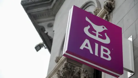 A sign for AIB outside a bank. It's purple with a white bird. The camera is looking up at the sign. The sky is overcast. The bank is a large building with decorative filigree around the windows and roof.
