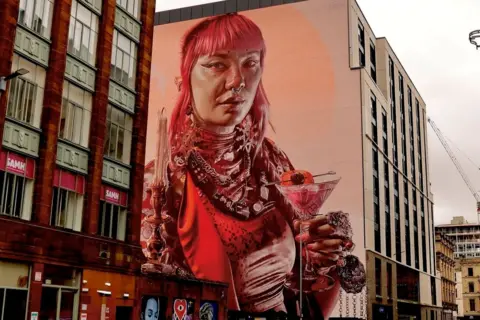 Graham Christie A large mural painted on a wall in a city street showing a woman with pink hair and red clothes holding a cocktail