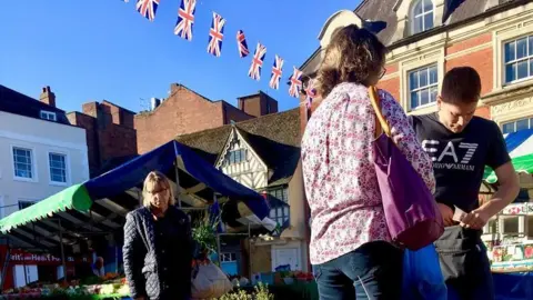 Cherwell District Council A market trader selling something to a woman whilst another looks on in Banbury.