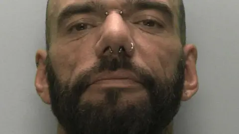 A mugshot of a man with a close crop, a dark beard and two rings through his nose looks into the camera