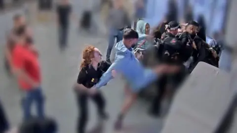 A man in blue kicks in the direction of a police officers while a police officer with red hair holds his arms behind him at the car park pay stations at Manchester Airport. 