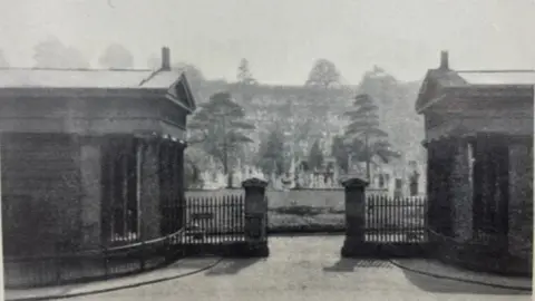 An historic image of the gates in black and white. The gates are central with the gatehouses either side and the cemetery behind, with trees and grass there.
At the front of the picture, it is paved.