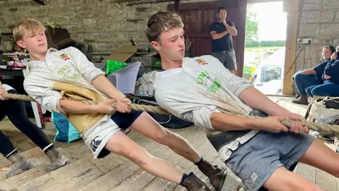 Two teenage boys pull on a tug of war rope. One boy has short blonde hair, wearing a white rugby shirt with various printed logos and a pair of white and black shorts. The other boy has short dark hair, is wearing an identical rugby shirt, black support belt and faded blue shorts. They are standing on a heavily grooved surface inside an untidy barn. A woman with short hair, a dark polo, jeans and boots is sat on the floor in the background. A man in a black t-shirt and shorts also stands in the background watching the pullers. A lorry can be seen through the open door
