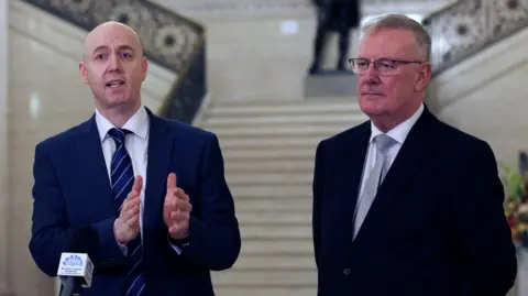 Jon Burrows standing beside Mike Nesbitt. Burrows is bald and is wearing a white shirt, navy tie and navy suit. Nesbitt has grey hair and i wearing a white shirt, grey tie and black suit. They are standing in front of the staircase in Stormont Buildings. 