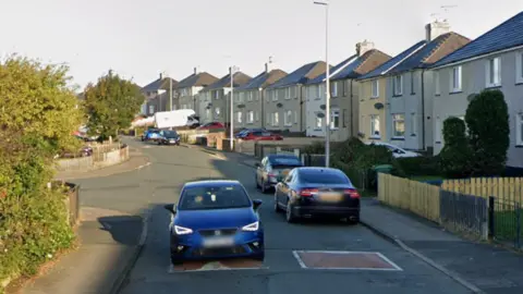 A Google Street View screenshot of Poole Road in Salterbeck, Cumbria. Semi-detached pebbledash properties line one side of the road, which has speedbumps, and several cars are parked next to the pavement. People's front gardens are visible on the other side of the road, at the edge of the photo.