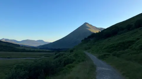 Neil Scotland The sun hits the top of a munro on a bright day, with a cycling path stretching along besides it 