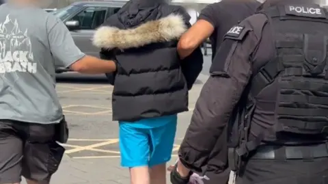 A man in a black duffel coat is led away by two men, while a man wearing police uniorm is seen in the foreground. 