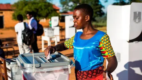 AFP via Getty Images A woman, in a patterned dress and satin blue vest, places her vote into a ballot box at an outdoor polling station. She has short hair.
