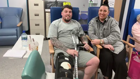 Nathan Davis has short brown hair and is wearing a brown T-shirt and shorts. He is on a chair holding crutches and his leg is tied. Her partner is sitting next to her and has black hair tied in a bun and is wearing a gray denim shirt and black trousers. 