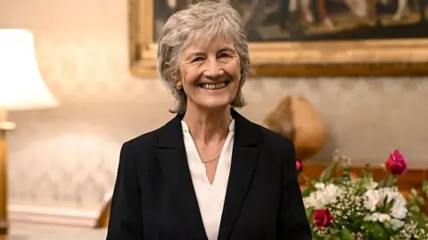 Getty Images Catherine Connolly smiling at the camera. She is wearing a white top, black blazer, gold earrings and a necklace. She has short grey hair. In the background, there are flowers, a painting and a lamp.