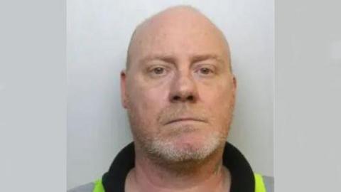 A police mugshot of Sean Begley. He is bald with a short white stubbly beard and standing against a grey wall.