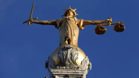 PA Media A golden statue of a person wearing a crown, holding a sword and a set of scales on top of a golden ball, which sits on a plinth.