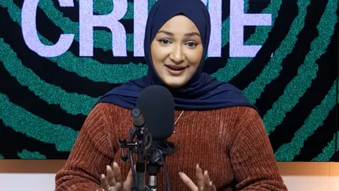 Rima Ahmed sits in front of a microphone, she wears an orange top and a dark blue headscarf