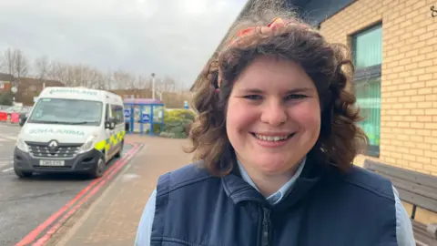 Lauren, who's wearing a navy blue porter's uniform, looking into the camera and smiling. She's got her curly brown hair held back by an orange patterned headband. On the left you can see an ambulance - Lauren is stood on the path leading to the hospital entrance. 