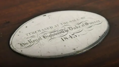 Woolley & Wallis An oval, silver plaque on top of a mahogany piece of wood. It is engraved with a line around its rim and writing which reads 'Purchase at the sale of His Royal Highness, The Duke of Sussex. 1843' written on it.