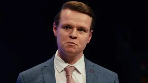 Getty Images Joshua Reynolds MP is pictured against a black background. He has short, neat brown hair and is wearing a blue suit with a pink tie. 