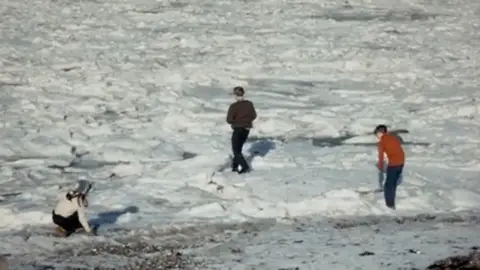 John Clague Three children stand in the frozen waves at the shoreline. One wears an orange jumper, another brown and the girl in a beige jumper. They walk on the snow which reaches up to the shore. 