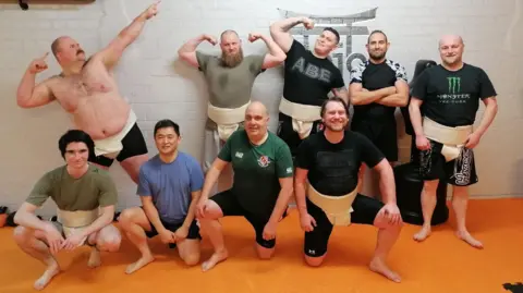 Barnsley Sumo Club A group of nine people posing together. They are standing and kneeling on an orange padded floor in front of a white brick wall that features a stylized graphic.
Most of the individuals are wearing mawashi over their training clothes. Several people in the back row are striking playful or flexing poses, while those in the front row are kneeling in a formation-like stance.