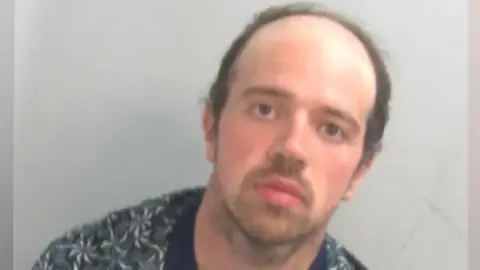 A police custody mugshot of Luke D'Wit, who is wearing an unbuttoned shirt with a T-shirt underneath. He has  short dark hair and a blank expression on his face.