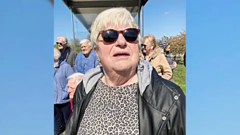 Jane Caines, wearing sunglasses, with other people at a bus stop in Ashton Vale in Bristol