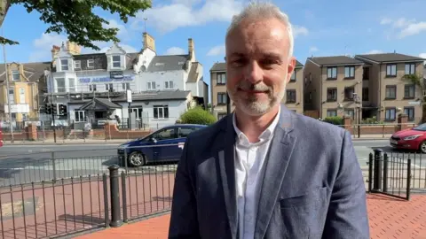 A man with grey hair and a grey beard wears a grey suit with a white unbuttoned shirt. He stands in front of the Albert Hotel on Anlaby Road. The white and grey building has scaffolding around it as it is currently being transformed into a House of Multiple Occupany after being given planning permission.