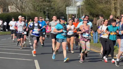 Crowds of people running along a road with spectators on the grass and pavement beside it.