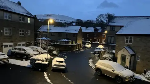 A street from above with cars, trees and rooftops covered in fresh snow. A mountain in the background is also covered in snow. 