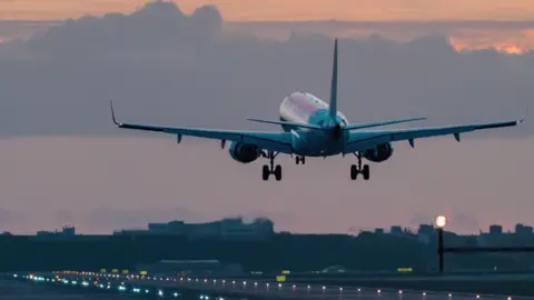 A plane taking off above a runway with the sun setting in the cloudy sky above it.