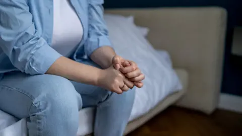 A woman wearing a blue shirt over a white top and blue jeans sits on the edge of a bed. She can only be seen from her shoulders down and has her hands folded.